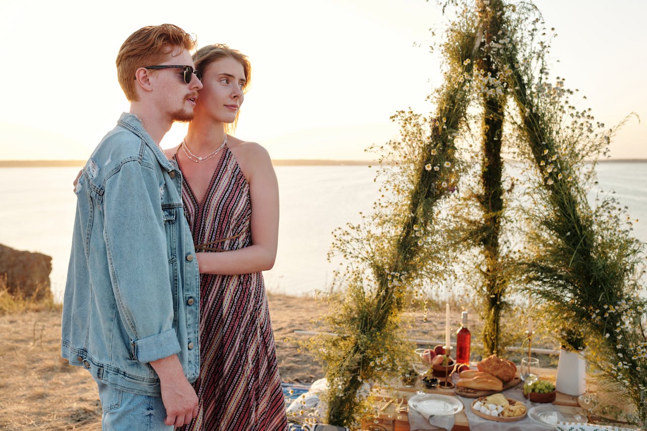 Couple embracing on a beachside picnic with food and floral decor at sunset.
