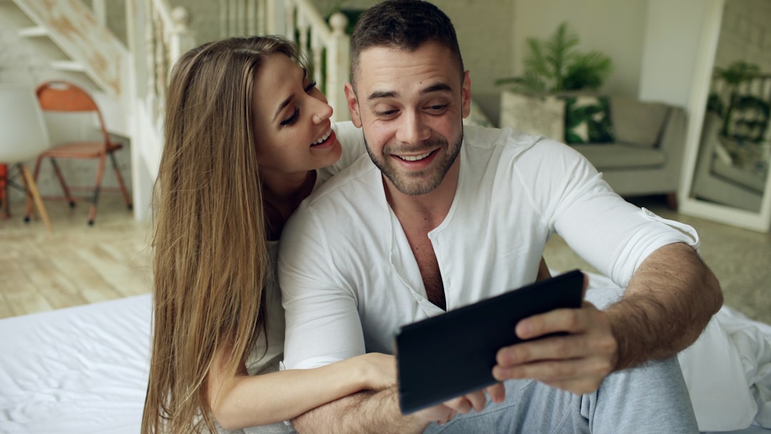 Young cute and loving couple having video chat holding tablet computer camera and chatting to friends sitting in bed at home